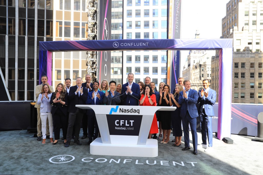 A large group of people stand in front of an arch and behind a NASDAQ desk