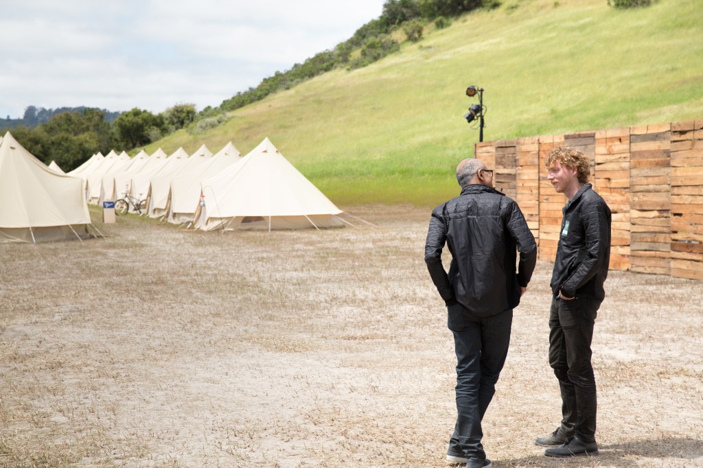 Two men stand near a row of tents talking